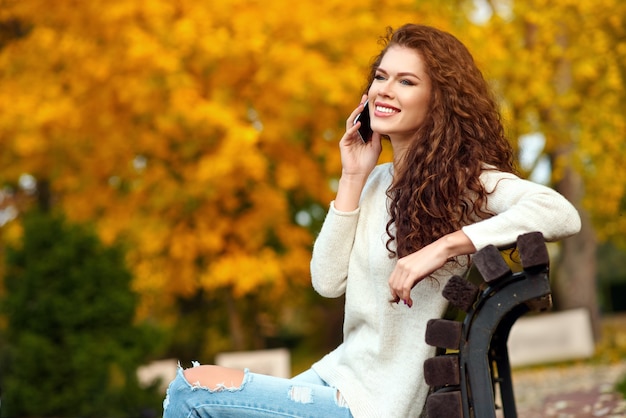 Junge Frau sitzt auf einer Bank im Herbst in einem Park und spricht auf einem Handy