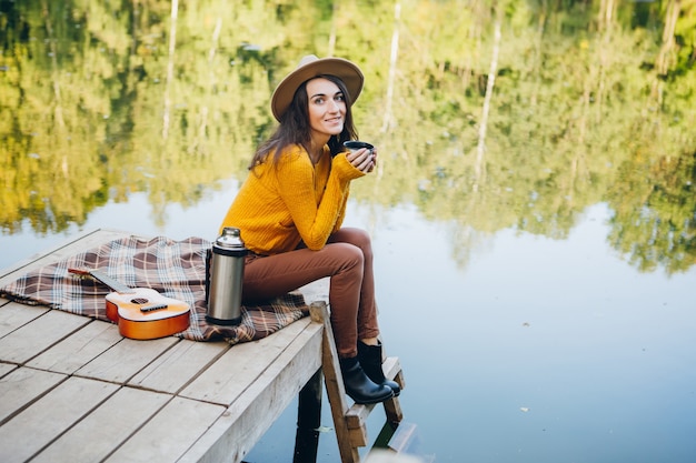 Junge Frau sitzt auf einer Brücke an einem See mit Herbstlandschaft und trinkt heißen Tee aus einer Thermoskanne. Tonisieren.