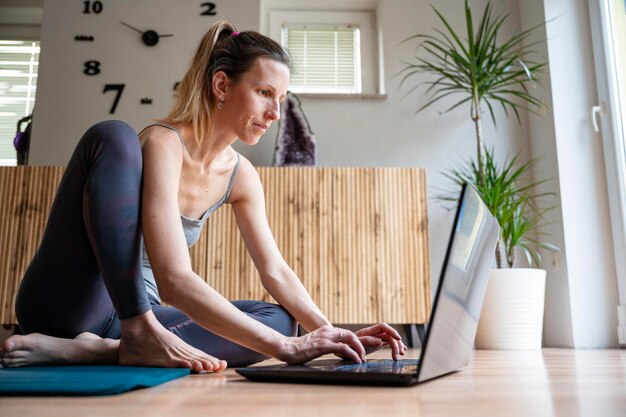 Junge Frau sitzt auf einer Matte auf dem Wohnzimmerboden und arbeitet oder studiert mit einem Laptop