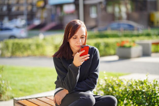 Junge Frau sitzt auf einer Parkbank und schaut auf ihr Handy Eine Frau mit Smartphone nutzt das Internet im Innenhof eines Wohnhauses