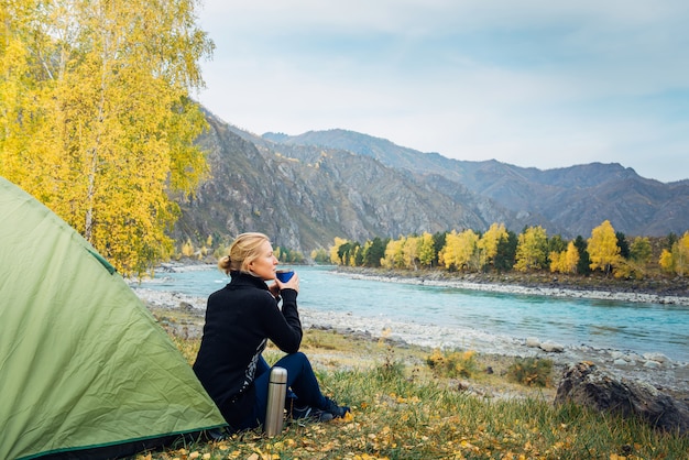 Junge frau sitzt auf gras nahe touristenzelt und trinkt heißen tee / kaffee von thermoskanne tasse im wald mit fluss und bergen.