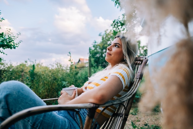 Foto junge frau sitzt auf holzstuhl in der nähe von van und trinkt tee im freien in der natur, während des sonnenuntergangs. sommer genießen, reisekonzept