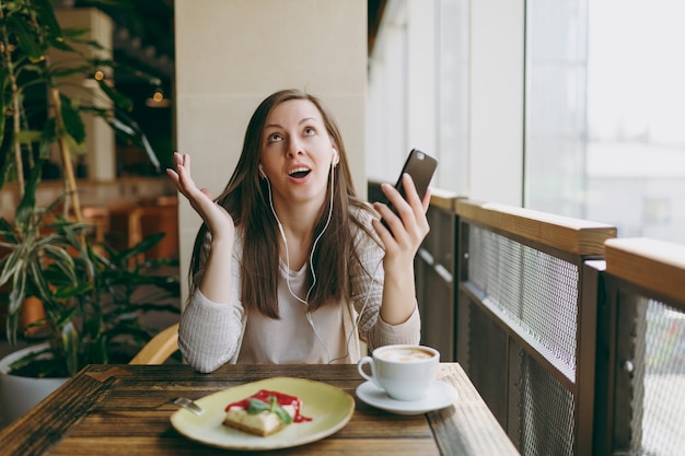 Junge Frau sitzt im Café am Tisch mit einer Tasse Cappuccino, Kuchen, entspannt sich in der Freizeit im Restaurant. Junge Frauen hören Musik in Kopfhörern auf dem Handy, ruhen sich im Café aus. Lifestyle-Konzept.