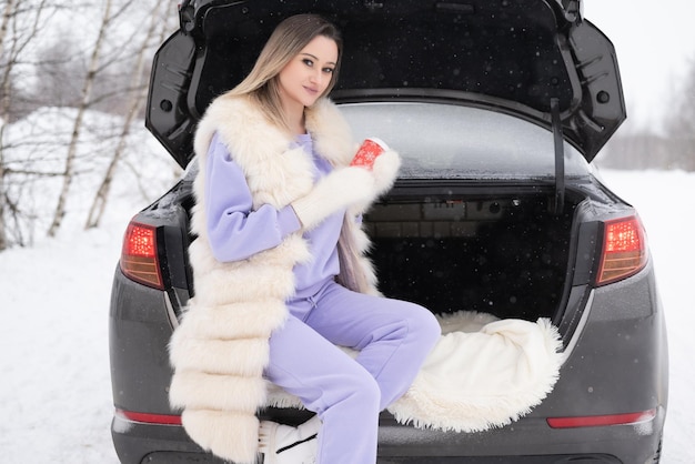 Junge Frau sitzt im Kofferraum des Autos und hält eine Tasse Heißgetränk in den Händen.