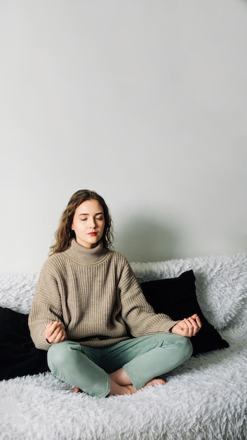 Junge Frau sitzt im Lotussitz auf dem Sofa, die Hände auf den Knien, und meditiert und versucht, ihren Geist zu entspannen und Stress abzubauen