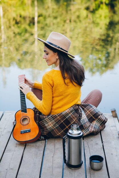 Junge Frau sitzt mit einer Gitarre auf einer Brücke auf einem See mit einer Herbstlandschaft. Tonisieren.