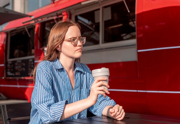 Junge Frau sitzt mit zusammenklappbarer Öko-Kaffeetasse in der Nähe von rotem Street Food Truck am sonnigen Sommertag a...