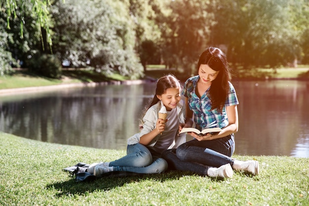 Junge Frau sitzt zusammen mit ihrer Tochter und liest ein Buch. Sie schauen sich gemeinsam ein Buch an. Mädchen lehnt sich an ihre Mutter. Sie isst Eis.