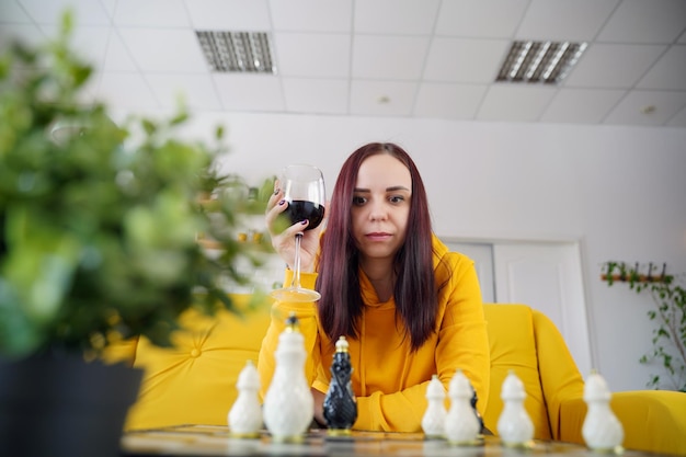 Junge Frau spielt Schach und trinkt Rotwein im Zimmer Erwachsene Frau mit Alkohol spielt in einem logischen Brettspiel, wobei sie selbst auf dem Sofa sitzt