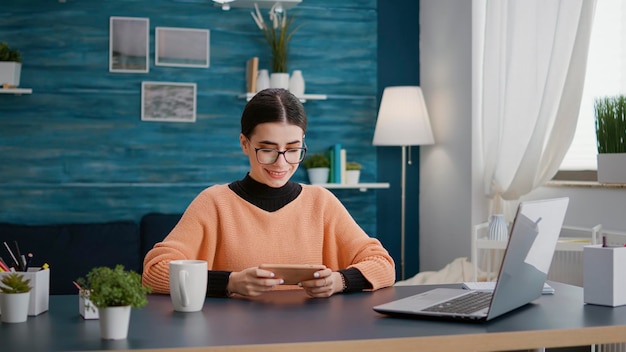 Junge Frau spielt Videospiele auf dem Smartphone am Schreibtisch und besucht die Schulklasse von zu Hause aus. Person, die ein Mobiltelefon verwendet, um ein Online-Spiel für Freizeitaktivitäten über eine Internetverbindung zu spielen.