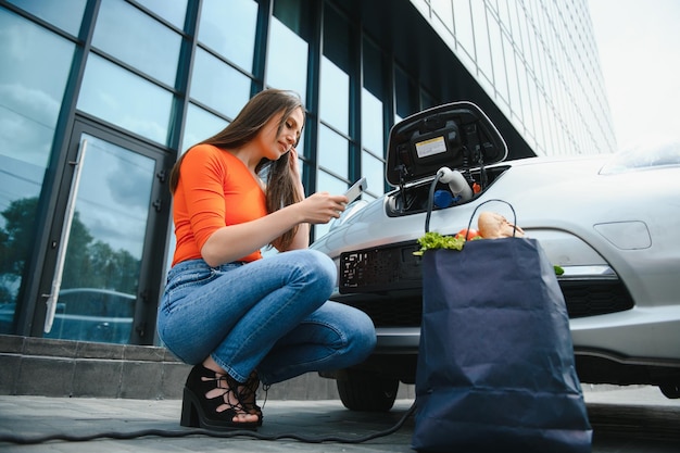 Junge Frau steht in der Nähe des Elektroautos und schaut auf das Smartphone Der Mietwagen lädt an der Ladestation für Elektrofahrzeuge Carsharing