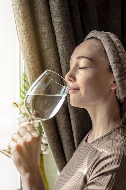 Junge Frau steht vor dem Fenster und genießt einen sonnigen Morgen und trinkt sauberes Wasser aus einem klaren Glas Konzept für einen angenehmen Start in den Tag und gesunde Gewohnheiten