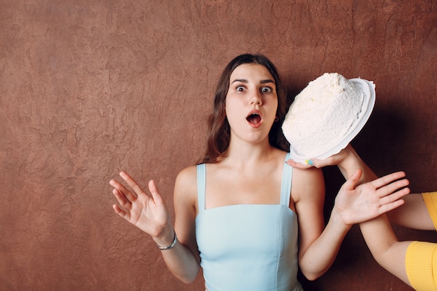 Junge Frau taucht Gesicht in weißen Kuchen mit Sahne auf braunem Wandhintergrund ein. Alles Gute zum Geburtstag Konzept.