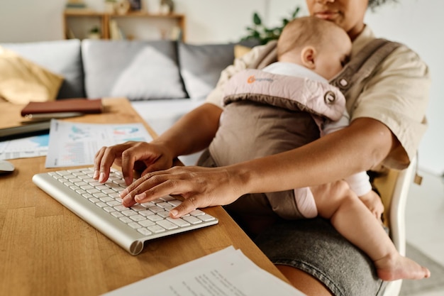 Junge Frau tippt auf der Tastatur, während ihr Baby im Tragetuch schläft und zu Hause arbeitet