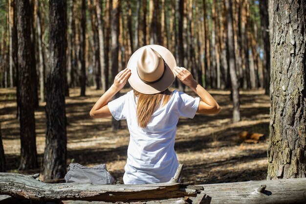 Junge Frau Tourist in Hut und T-Shirt sitzt auf einem Baumstamm während eines Halts im Wald. Rückansicht.