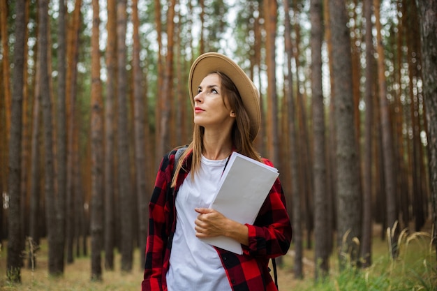 Junge Frau Touristin in einem Hut, rotes kariertes Hemd hält eine Karte des Gebiets im Wald.