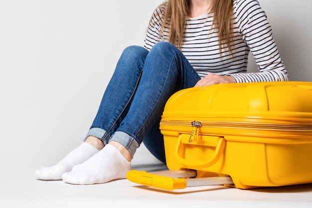 Junge Frau Touristin in Jeans sitzt in der Nähe eines gelben Koffers auf einem hellen Hintergrund