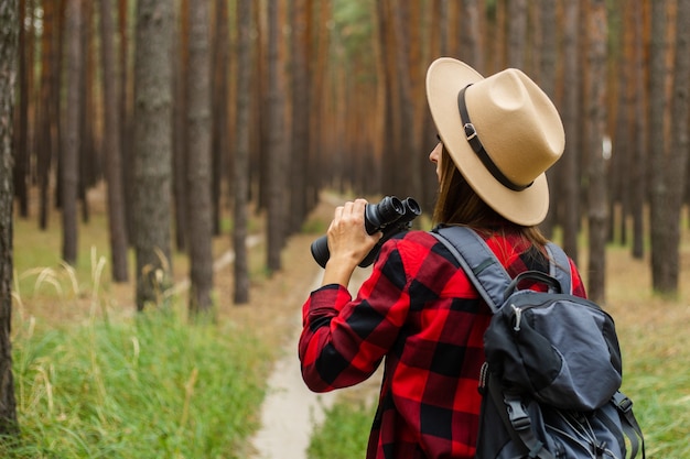 Junge Frau Touristin mit Rucksack, Hut und rotem kariertem Hemd und schaut durch Fernglas im Wald.