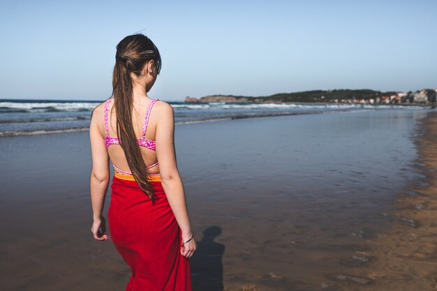 Junge Frau trägt ein rotes Handtuch und einen rosa Bikini zu Fuß am Wasserrand am Strand