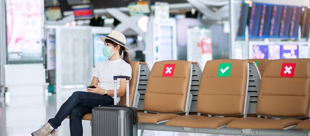 Junge Frau trägt Gesichtsmaske und sitzt auf Stuhl im Flughafen, Schutz Coronavirus-Krankheit (Covid-19) -Infektion, asiatische Reisende. New Normal, Reiseblase und soziale Distanzierung