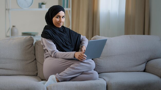 Junge Frau trägt Hijab, sitzt auf dem Sofa im Wohnzimmer, bewegt die Hände auf dem Laptop und genießt die Arbeit von zu Hause aus