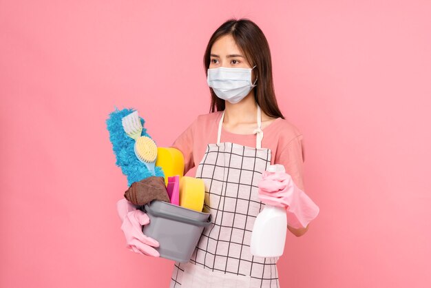 Junge Frau trägt Masken, um Krankheiten und Schürzen zu schützen, indem sie einen Eimer mit Reinigungsmitteln auf rosafarbenem Hintergrund hält.