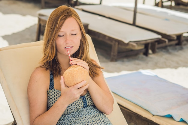 Junge Frau trinkt Kokosmilch auf Chaiselongue am Strand.