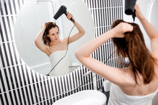 Junge Frau trocknet und stylt Haare mit Fön und macht Frisur im modernen Badezimmerinnenraum Beauty-Routine nach der morgendlichen Dusche