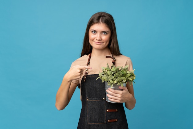 Junge Frau über dem lokalisierten Blau, das einen Blumentopf nimmt und ihn zeigt