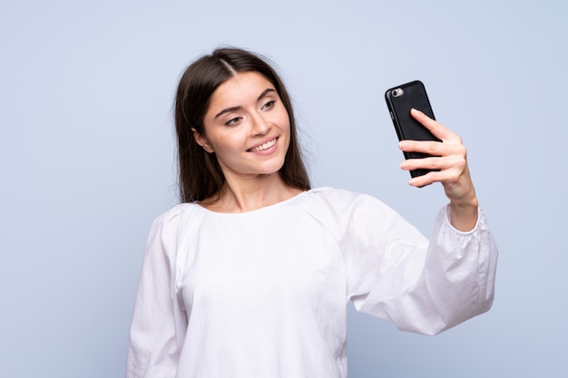 Junge Frau über der lokalisierten blauen Wand, die ein selfie mit dem Mobile nimmt