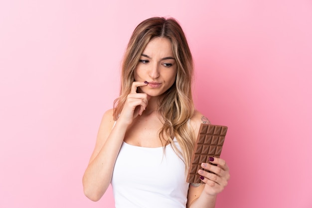 Junge Frau über der lokalisierten rosa Wand, die eine Schokoladentablette nimmt und Zweifel hat