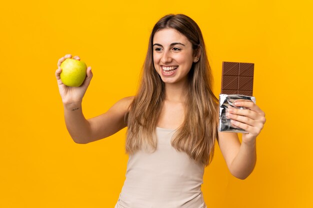 Junge Frau über isolierte gelbe Wand, die eine Schokoladentafel in einer Hand und einen Apfel in der anderen nimmt