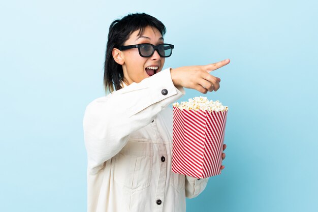 Junge Frau über isoliertem Blau mit 3D-Brille und hält einen großen Eimer Popcorn, während weg zeigend