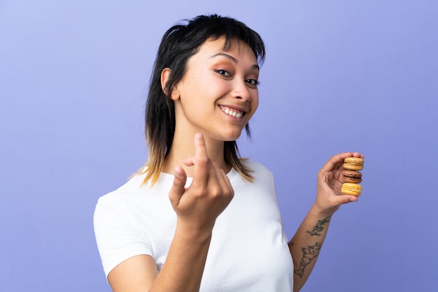 Junge Frau über isoliertem Purpur, das bunte französische Macarons hält und einlädt, zu kommen