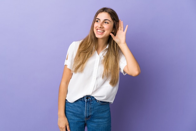 Junge Frau über lokalisiertem lila Hintergrund, der etwas hört, indem man Hand auf das Ohr legt