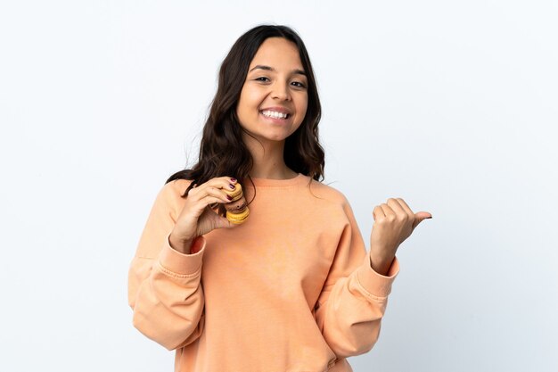 Junge Frau über lokalisiertem weißem Hintergrund, der bunte französische Macarons hält und Seite zeigt