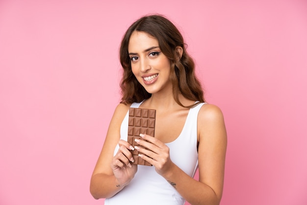 Junge Frau über rosa Wand, die eine Schokoladentafel nimmt und glücklich