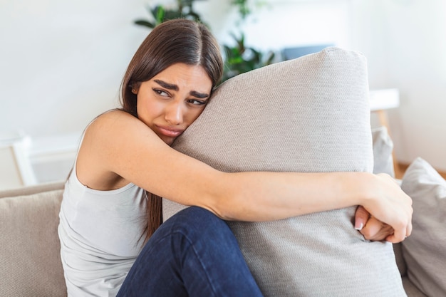 Junge Frau umarmt Kissen mit Depressionen und weint auf dem Sofa zu Hause. Einsame Millennial-Dame, die sich gestresst und hoffnungslos fühlt, an Stimmungsstörungen leidet oder psychische Probleme hat