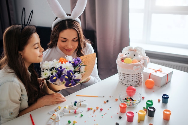 Junge Frau und ihre Tochter bereiten sich auf Ostern vor. Sie riechen zusammen Blumen. Malen Sie mit Süßigkeiten und Dekoration auf dem Tisch. Tageslicht.
