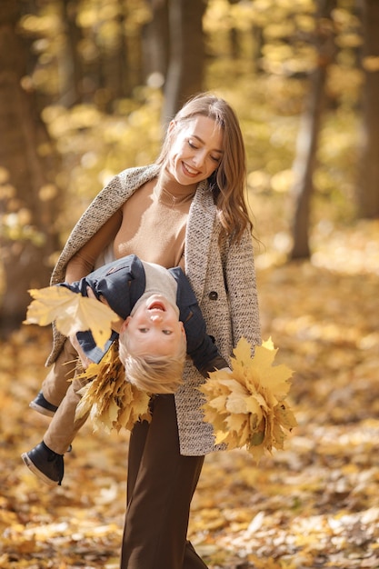 Junge Frau und kleiner Junge im herbstlichen Wald. Frau, die ihren Sohn auf ihren Händen hält. Junge, der modische Kleidung trägt und gelbe Blätter hält.