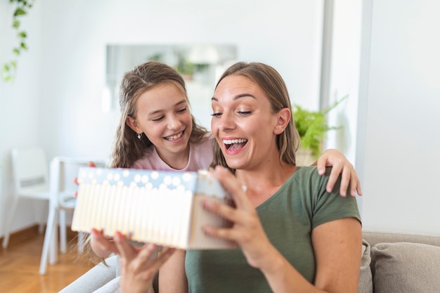 Junge Frau und Mädchen zu Hause feiern den Muttertag sitzen auf dem Sofa Tochter umarmt Mutter küssen Wange Mutter lachen freudig halten Geschenkbox