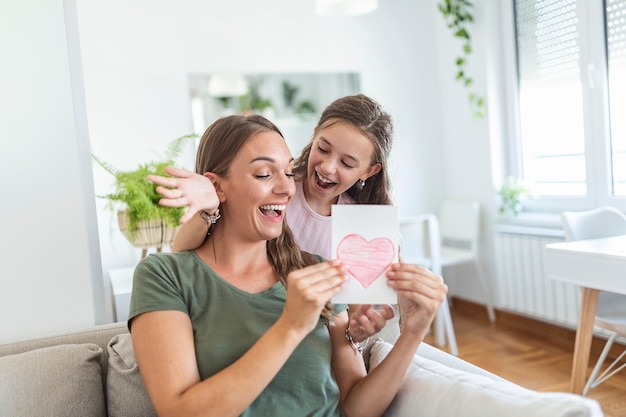 Junge Frau und Mädchen zu Hause feiern den Muttertag sitzen auf dem Sofa Tochter umarmt Mutter küssen Wange Mutter lachen freudig halten Geschenkbox