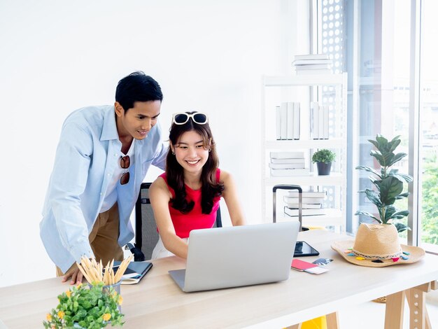 Junge Frau und Mann des glücklichen asiatischen Paares, die zusammen Laptop-Computer auf Schreibtisch verwenden, um Flugbuchungshotelzimmer und Reiseinformationen im Büro zu reisen, bereit, glückliches Feiertags-Sommerferienkonzept zu reisen