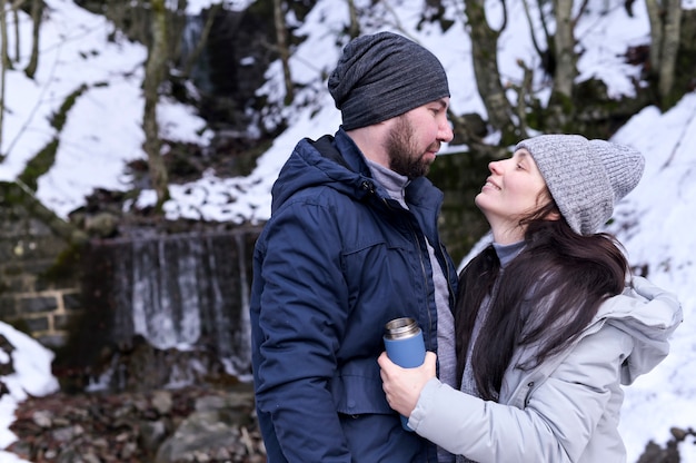 Junge Frau und Mann, die in einem verschneiten Wald kuscheln. Ein Paar Liebhaber in Winterkleidung. Das Mädchen hält eine Thermoskanne in den Händen. Speicherplatz kopieren