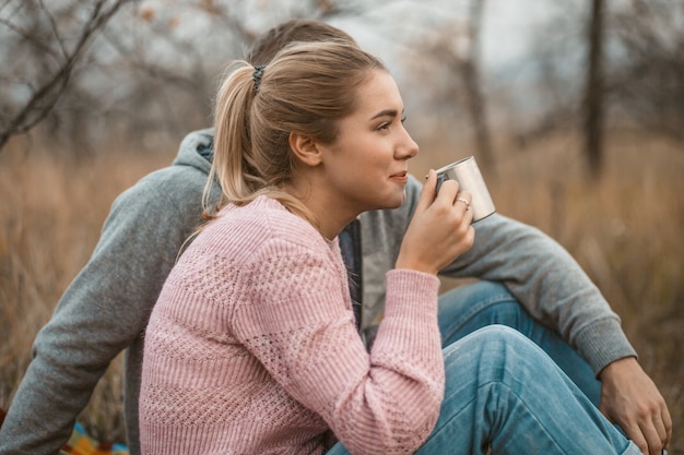 Junge Frau und Mann ruhen, heißen heißen Kaffee oder Tee trinkend, während sie auf Decke im Herbstgras draußen sitzen.