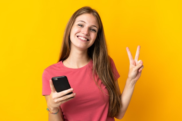 Junge Frau unter Verwendung des Mobiltelefons lokalisiert auf gelbem Hintergrund lächelnd und Siegeszeichen zeigend