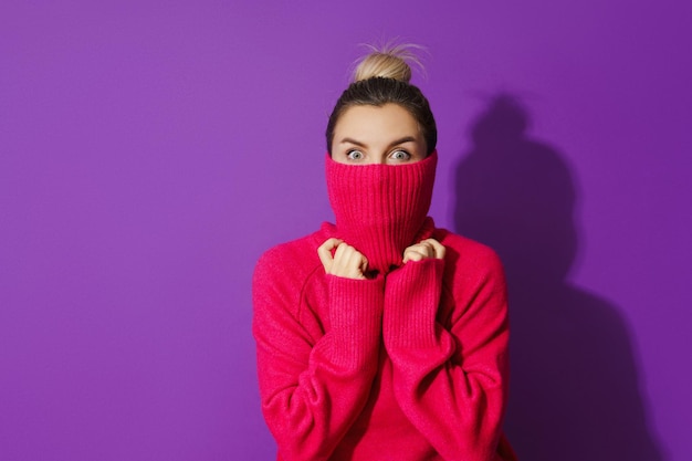 Junge Frau versteckt ihr Gesicht in einem warmen und gemütlichen Pullover mit Rollkragen vor violettem Hintergrund