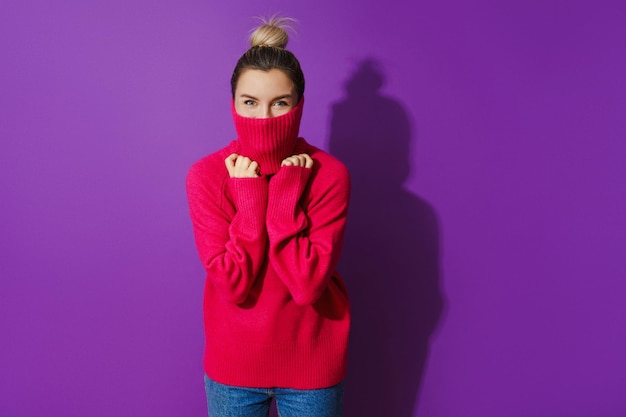 Junge Frau versteckt ihr Gesicht in einem warmen und gemütlichen Pullover mit Rollkragen vor violettem Hintergrund