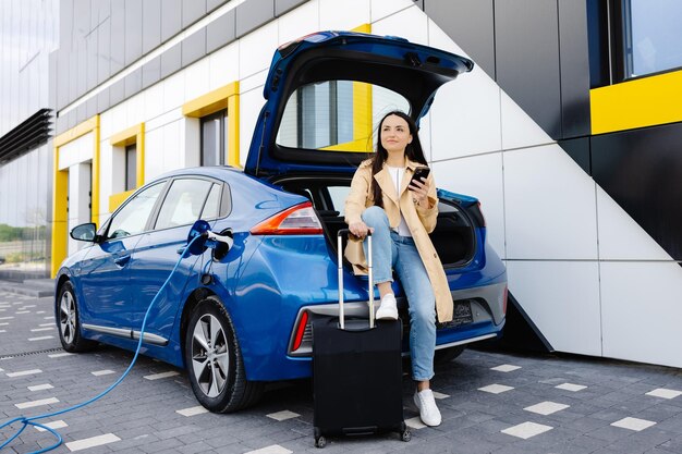 Junge Frau wartet darauf, die Autobatterie von einer kleinen öffentlichen Station aufzuladen und nutzt ihr Smartphone, während sie das Auto auflädt