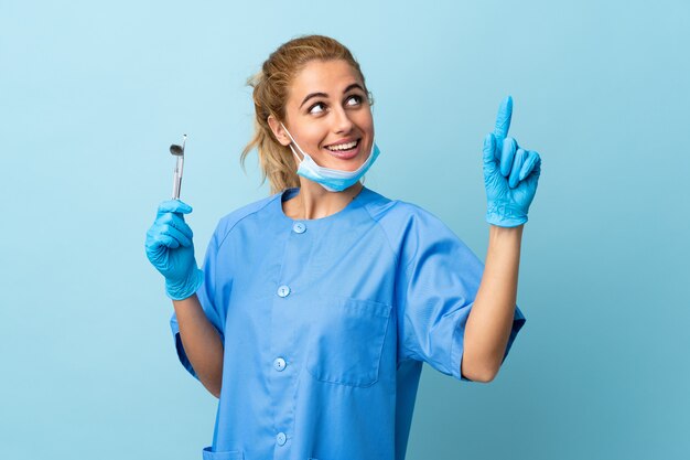 Junge Frau Zahnarzt, die Werkzeuge auf lokalisiertem blauem Zeigen mit dem Zeigefinger eine große Idee hält